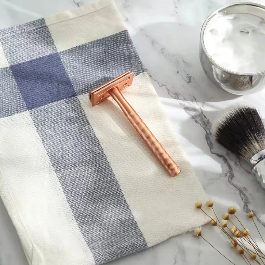 Rose gold safety razor on a checkered towel with a brush and jar on a marble surface
